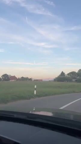 The video shows a point-of-view shot from a vehicle driving along a rural road. The sky is a pale blue with wispy clouds. In the distance, there are rolling green fields, trees, and houses. Mountains are visible on the horizon. The road is paved with a white line marking the edge. There are a few traffic signs and poles visible along the roadside. The sun appears to be low in the sky, casting a soft light.