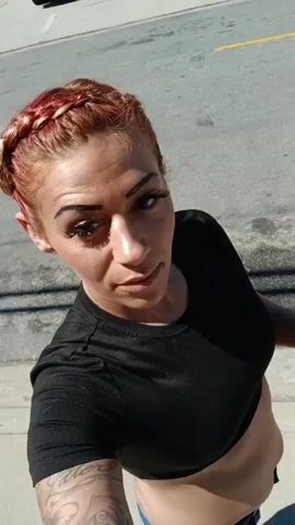 A woman with red braided hair, wearing a black crop top and blue jeans, stands outside and holds a cigarette. She looks at the camera while holding the cigarette, then lowers the camera to capture the cigarette in her hand and her torso with the crop top ending above the jeans. She then pans the camera downwards to show her legs, white sneakers, and shadow on the sidewalk. She lifts the camera slightly to show the back of a silver car next to her.