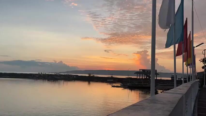 The video opens with a wide shot of a tranquil body of water reflecting the soft hues of a sunrise. The sky is painted with shades of orange, pink, and yellow, with wispy clouds adding texture. A distant landmass is visible on the horizon. In the foreground, a white railing and sidewalk run along the right side of the frame. Several flagpoles stand along the sidewalk, each adorned with colorful flags that flutter gently in the breeze. The flags are white, light blue, red, and orange. The water is calm, with small ripples indicating a light breeze. Along the left side of the water, a long, narrow pier or breakwater extends into the distance, lined with several small fishing boats. Some structures are visible on the pier, possibly shelters or small buildings. The overall atmosphere is peaceful and serene, capturing the quiet beauty of a coastal sunrise. There are no people visible in the shot.