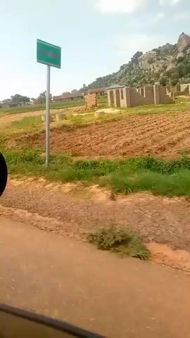 The video shows a moving vehicle's perspective of a rural landscape. The scene unfolds from a vehicle, likely a car, with the side mirror visible on the left. The road appears to be dirt or gravel, with reddish-brown soil. The landscape features cultivated fields, some appearing bare and others with rows of crops or vegetation. In the background, there are several buildings, likely houses or small structures, some with corrugated metal roofs. A prominent rocky hill or mountain range is visible in the distance. A utility pole with wires is also present. A distinct white and black striped marker or signpost is seen on the side of the road. Trees are scattered throughout the scene, with one larger tree standing out in the latter part of the video. The sky is partly cloudy. The overall impression is of a sunny day in a developing rural area. No people are clearly visible in the video.