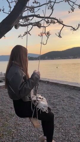 A young woman with long brown hair is sitting on a swing by the water. She is wearing a dark jacket, black pants, and light-colored shoes, and has a white bag slung over her shoulder. The swing is suspended from a tree branch. The water is calm and reflects the colors of the sunset. In the background, there are hills and a town. The ground under the swing is covered in small grey stones.