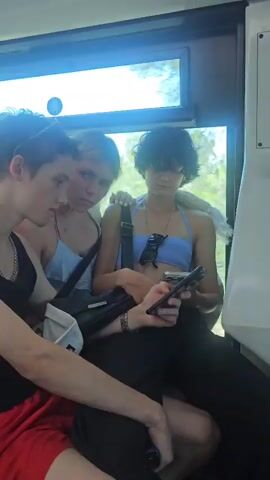 Three young people, two females and one male, are seated on a vehicle, possibly a train or bus. The male, on the left, wears a black tank top and a red garment around his waist. He is looking at the female next to him. The female in the middle has blonde hair and wears a light blue top. She is looking at the male. The third person, with dark curly hair, wears a light blue halter top and dark pants. They are seated on the right, looking at a smartphone in their hands. A black bag is placed between the middle and right individuals. The environment appears to be the interior of a moving vehicle, with a window visible in the background showing a bright, outdoor scene.