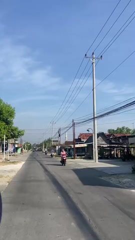 The video shows a street scene with a clear blue sky and a few clouds. Several utility poles with numerous overhead wires are prominent. Buildings line the right side of the street, with various roofs visible. On the left side of the street, there are more buildings and some trees. Several motorcycles are present: one is being ridden by a person in a pink headscarf and light-colored clothing moving from right to left in the foreground. Another motorcycle is being ridden by a person in a light blue shirt from left to right further down the road. A third motorcycle is visible further in the distance, also moving from left to right. A fourth motorcycle is parked on the right side of the street. The road is paved and appears to be in good condition, with tire marks visible. The overall environment suggests a rural or semi-urban setting.