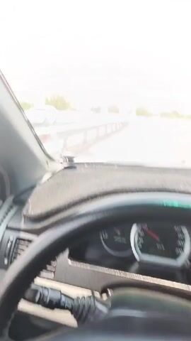 The video shows a first-person perspective from the driver's seat of a car moving on a highway. The dashboard and steering wheel are visible in the foreground. The road ahead is clear with a concrete barrier on the left and trees and hills in the background. A white truck is seen on the opposite side of the highway. The sky is bright and appears to be a sunny day.