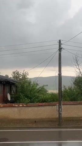A static wide shot captures a rainy day in a rural area. In the foreground, a wet asphalt road stretches across the bottom of the frame. Above the road, a low brick wall with a textured beige facade runs horizontally. Behind the wall, a dense line of green bushes and trees partially obscures a distant landscape of rolling hills under a cloudy, overcast sky. To the left, a portion of a house with a dark roof and a satellite dish on its side is visible. A tall, dark utility pole with multiple power lines extending across the frame stands prominently on the right side of the road. Rain can be seen falling steadily throughout the video.