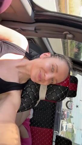 A woman with tied-back hair, a nose ring, and wearing a black tank top is in the back of a vehicle. A child in a pink shirt lies asleep. A man in a striped shirt sits beside the child. The car has black and red diamond-patterned seat covers. Other cars are visible outside.
