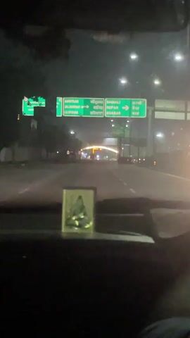 The video is filmed from inside a vehicle at night. The perspective is looking forward through the windshield. A green highway sign is visible overhead, displaying directions to Amritsar, Jalandhar, Ludhiana, Manali, Ropar, and Kharar, with arrows indicating the directions. The road ahead is paved and illuminated by streetlights. In the distance, a structure with illuminated arches is visible. The interior of the car is dark, but a small decorative item is placed on the dashboard. The lighting is dim, suggesting it is nighttime or a poorly lit area. The weather appears foggy or misty, as indicated by the haziness of the light in the distance. There is a person's leg visible in the lower right corner of the frame. The overall environment appears to be a highway or a major road on the outskirts of a city.