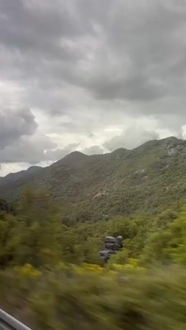 The video shows a view of mountains covered in green vegetation under a cloudy, overcast sky. The perspective appears to be from a moving vehicle, with blurred greenery in the foreground. The mountains recede into the distance, and the sky is filled with gray clouds.