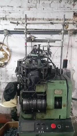 The video displays a knitting machine in operation within an industrial setting, likely a workshop or factory. The machine is predominantly green with intricate metal components and wires. A black fabric drapes over a part of the machine, and a spool of black thread is visible. The background features a rough, white brick wall with some weathered, possibly decorative, elements on the left. The floor appears to be concrete, with a brown plastic bin containing dark blue material at the bottom of the frame. A prominent red button is located on the control panel of the machine. The lighting suggests an indoor environment, possibly with artificial light given the consistent brightness across the frame. No people are visible. The overall impression is of a functional industrial space where textile production is taking place.