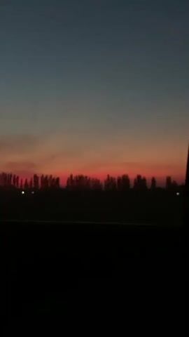 This is a video of a sunset viewed from inside a vehicle. The sky is a gradient of blue and orange. Silhouetted trees form a line across the horizon. The interior of the vehicle is dark, obscuring the front.