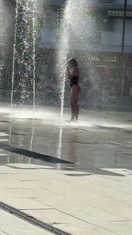 A person in a black swimsuit stands in a shallow water fountain on a paved surface. Water sprays up around them from multiple jets, creating a mist. The person is looking up and appears to be wiping their face or hair. In the background, there are buildings, a motorcycle, and other people. The sun is shining, casting reflections on the wet ground.