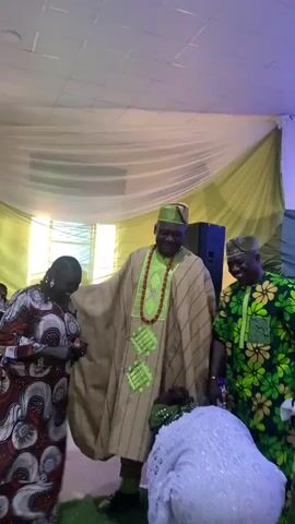 Here's a description of the video:

The video shows three individuals in what appears to be an indoor event space. A man in the center wears a light brown and green striped robe, a green hat, and a red beaded necklace. He smiles and briefly adjusts his robe. To his left is a woman in a red patterned dress, and to his right is another man wearing a green and yellow patterned outfit with a similar hat.  The background is decorated with white and green drapes, suggesting a festive occasion. A black speaker is visible behind the man in the center.  There seems to be some sort of celebration or gathering taking place.