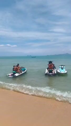 Two jet skis are in the shallow water near a beach. One jet ski has two people on it, and another jet ski has one person on it. There is another jet ski to the right of the one with one person. The sky is blue with some clouds. The water is clear and calm. The beach is sandy.