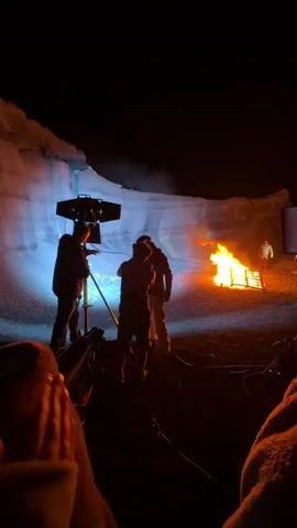 The video shows a film set at night with a large bonfire. A crew of at least four people are standing near a film light, while a fire blazes on the snowy ground. A white wall is visible behind the fire, illuminated by the light. There appears to be one person in a lighter colored shirt walking in the background, past the fire. The camera appears to be located behind someone wrapped in a fuzzy blanket. The scene suggests they are filming a scene involving a fire in a cold, possibly wintery, environment.