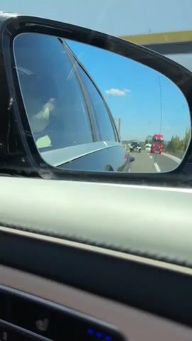 This video shows a reflection in a car's side mirror. The reflection shows traffic on a highway under a clear blue sky. There are several vehicles, including cars and trucks. The side mirror is positioned to the left of the frame, with the interior of the car visible in the foreground. The video appears to be captured while the car is in motion.