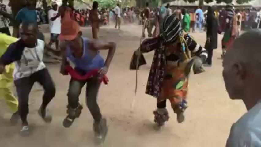 The video shows a group of dancers in an outdoor setting. The dancers are wearing traditional clothing and appear to be performing a cultural dance. One dancer is wearing a mask and colorful robe and using a walking stick. They are surrounded by a crowd of people watching the performance. The ground is dusty, and there are trees in the background.
