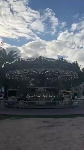 The video shows a carousel in operation.  It has a dark color scheme and is adorned with lights, and a decorative edge. A fence surrounds the carousel. The ground around it is a mix of sand and grass. Behind the carousel, there are trees and buildings. The sky is blue with white clouds.