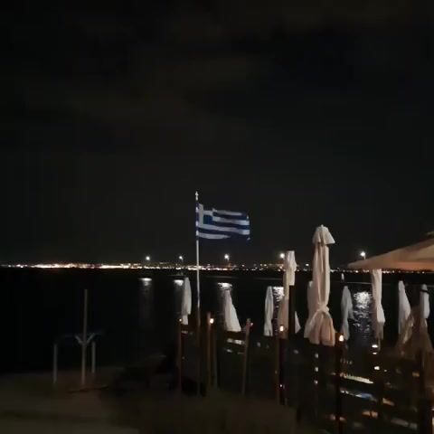 The media shows a nighttime scene on a beach or waterfront area in Perea, Greece. A Greek flag is prominently displayed on a flagpole, waving gently. In the background, the calm sea reflects distant lights, suggesting a town or city across the water. The foreground features a beachside establishment, likely a restaurant or bar, with numerous closed white umbrellas and some seating areas. The area is dimly lit by small lamps or candles placed on tables, creating a cozy and serene ambiance. The sky is dark with some visible clouds, indicating it is nighttime. A few people are faintly visible in the background, seated at tables under one of the awnings, enjoying the evening. The overall mood is peaceful and relaxing, typical of a seaside evening setting. A tall, feather-shaped flag with the text "Dicristo" and a purple logo is visible on the right side of the frame.