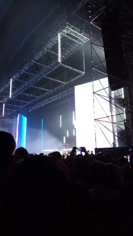 A concert crowd is silhouetted in the foreground, facing a stage. The stage is lit with bright white and blue lights, creating a hazy effect. A drum set is visible on stage. People in the crowd are holding up their phones, seemingly recording the performance. Overhead, a complex grid of metal beams supports the stage lighting.