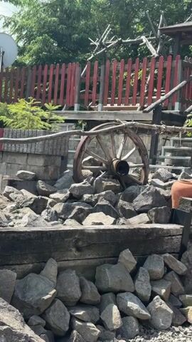 The video depicts an outdoor scene, likely a garden or terraced yard in Wisła, Poland, under bright, sunny conditions. The foreground features a substantial collection of light grey rocks forming a base, topped by a dark wooden beam. More rocks are piled behind this, creating different levels within the landscape.

The primary subject is a bright pink foam toy airplane, speckled with blue dots. At the beginning of the video, a hand briefly appears, holding and then throwing the plane. The plane glides through the air, briefly flying high above the immediate foreground.

As the plane flies and the camera pans, more of the setting is revealed. A large, weathered wooden wagon wheel is prominent among the rocks, leaning against a stone and wood structure. Nearby, a terracotta pot rests on the rocks. In the background, a distinct red wooden picket fence runs horizontally across the scene. Behind and around the fence, numerous green trees and smaller bushes contribute to a lush, natural environment. A white satellite dish is visible above the fence line.

The pink toy plane descends and lands gently among the rocks near the wooden wheel and terracotta pot. No discernible text is visible in the video. The activity appears to be a casual, recreational outdoor moment.