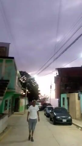 A man in a white t-shirt and gray shorts walks down a street at dusk. He is walking towards the camera. The street is lined with buildings and parked cars. The sky is cloudy and purple. There are power lines running overhead.