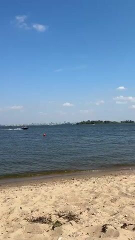 A powerboat with at least two occupants wearing life vests speeds across a wide body of water. The boat leaves a white wake as it moves from left to right across the frame. In the foreground, a sandy beach is visible. In the mid-ground, an orange buoy floats on the water. In the background, a distant city skyline with buildings of varying heights is visible, interspersed with trees and vegetation. The sky above is blue with scattered clouds.