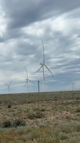 The video shows a field of wind turbines under a cloudy sky. The wind turbines are tall and white with large blades that are spinning. The ground is covered with dry grass and shrubs. There are also a few utility poles scattered throughout the landscape. The video is shot from a moving vehicle, as evidenced by the slight blur of the foreground. The overall impression is one of a vast, open landscape, possibly in a rural or industrial area.
