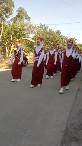 The video shows a group of young girls, likely students, dressed in matching red uniforms with white hijabs and decorative headwear resembling feathers. They appear to be participating in some sort of procession or parade, as they are lined up on a paved road. Some of the girls have bibs with numbers on them, and one bib clearly displays "064". They are all facing towards the viewer and seem to be in formation. The background consists of trees and a clear sky, suggesting an outdoor setting during the daytime. There is also a motorcycle visible in the lower left portion of the frame. The camera perspective is low and angled, making the video appear tilted.