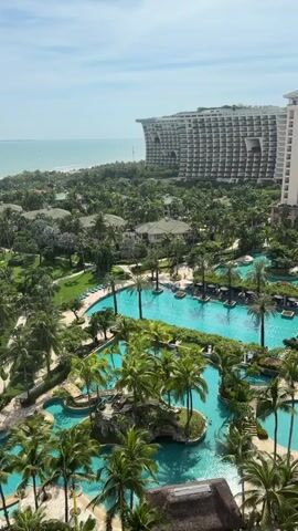 This video shows an aerial view of a resort on a sunny day. The resort features numerous swimming pools with blue water, surrounded by lush palm trees and green landscaping. In the background, there is a beach with white sand and the turquoise ocean stretching out to the horizon. A large, modern hotel with a distinctive curved facade is visible on the right side of the frame, overlooking the resort and the sea. The sky is blue with scattered white clouds.