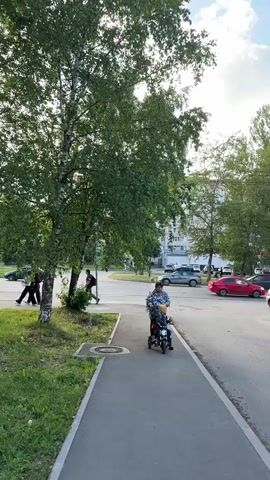 The video shows a suburban street scene in Vologda, Russia, during the daytime. The sun is shining brightly, casting dappled shadows through the lush green leaves of the birch trees lining the sidewalk. A woman is pushing a child on a small, red tricycle along the paved walkway. Further down the street, several people are walking, and cars are driving by. One car, a red sedan, is visible on the right side of the road.

In the background, a multi-story building can be seen, suggesting a residential area. There's also a large billboard visible, displaying what appears to be Russian text and an image of greenery. The overall atmosphere seems peaceful and ordinary, with the subjects engaged in everyday activities like a stroll in the park or a bike ride. The weather appears to be pleasant, with a few white clouds dotting the blue sky. The time of day is likely late afternoon or early evening, given the angle of the sunlight.