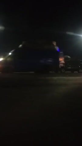 The video shows a dark, nighttime scene. A truck with its headlights on is visible, carrying a large load of what appears to be hay or straw. There are other vehicles and structures in the background, illuminated by streetlights. A person walks past the frame in the foreground at one point. The scene suggests a roadside or parking area at night, possibly a transportation hub or a place where goods are being loaded or unloaded. The lighting is dim, and the overall atmosphere is somewhat obscured by darkness.