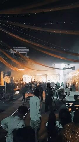 The video shows a nighttime outdoor film set. There are a number of people milling around, including camera operators and other crew members. Several cameras on tripods are pointed towards a central area behind a set of black metal frames. There is a bright light source in the central area, partially obscured by smoke or haze. Overhead, strands of orange fabric and fairy lights are strung across the set. Two white buildings are visible in the background.