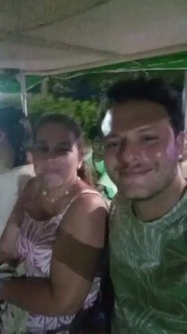 The video shows a selfie taken by a man in the foreground, smiling at the camera. To his left, a woman is also smiling. Both are seated, and the background is dark, suggesting an evening or nighttime setting. There are hints of green foliage and possibly some lighting fixtures above. Another person is partially visible to the far left, also appearing to be seated. The man is wearing a green shirt with a leaf pattern, and the woman is wearing a patterned dress. The overall impression is of a casual, social gathering, possibly outdoors.