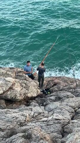 Two men are standing on rocky terrain near a body of water. The man on the left is wearing a blue t-shirt and dark pants. He is sitting on a rock and gesturing with his left hand towards the right. The man on the right is wearing a dark t-shirt and dark pants. He is standing and holding a long fishing rod, which is extended over the water. Another fishing rod is visible on the right side of the frame, stuck into the rocks. A black bag is on the ground between the two men. The water is a vibrant green color and appears to be choppy, with waves breaking against the shore. The rocks are rough and uneven.