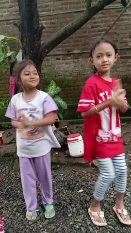 Two young girls are dancing in a yard. The girl on the left is wearing a white t-shirt with a cartoon character, lavender pants, and sandals. The girl on the right is wearing a red t-shirt, striped leggings, and sandals. They are both smiling and moving their arms and bodies to the music. The yard is covered in small stones, and there is a tree, a brick wall, and some plants in the background. There is also a white and red cooler visible in the background.