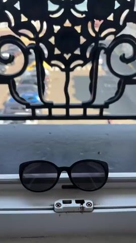 The video begins with a close-up of sunglasses resting on a ledge. The camera then pans up and out to reveal a view from a balcony or window. In the foreground, there is a decorative black metal railing with a heart-shaped design. Beyond the railing, a Ferris wheel is prominently displayed against a bright blue sky and the ocean. A beach area and a parking lot filled with cars are visible below and in front of the Ferris wheel. The overall scene depicts a sunny day at a seaside location.