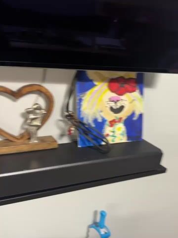 The camera pans left across a shelf. On the shelf are various decorative items, including wooden letters spelling "HOME," a wooden heart-shaped ornament with a metal figurine inside, and a child's drawing of a person wearing a black suit with the word "Nike" on the shirt and blue boots. A black television screen is visible above the shelf. The background is blurred and out of focus.