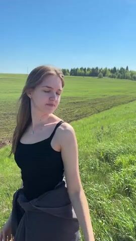 A young woman stands facing away from the camera in a grassy area. She is wearing a black tank top, and has a gray sweatshirt tied around her waist. Her brown hair is blowing in the wind.  Beyond her, a large field stretches to the horizon under a clear blue sky. An oil pump is visible in the distance. The field is partly green with crops, and partly brown with bare earth and tire tracks.