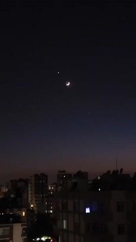 The video captures a twilight sky above a city skyline. The sky is a deep purple transitioning to a lighter shade near the horizon. Three celestial bodies are visible: a crescent moon, and two bright stars or planets. Below the sky, the silhouettes of various buildings are visible, some with illuminated windows. The overall atmosphere is calm and serene, suggesting an evening scene.