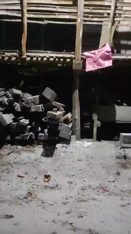 The video shows an outdoor scene at night. There is a pile of bricks to the left of the frame, and a wooden structure with horizontal planks overhead. A piece of pink fabric is hanging from a wooden post on the right side of the structure. The ground is covered in dirt and debris.
