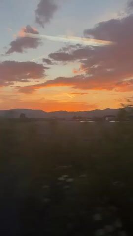 The camera, positioned inside a vehicle, possibly a train or car, captures a landscape moving past during sunset. The sky is streaked with orange and pink clouds against a light blue backdrop, with visible contrails from airplanes. The setting sun is situated behind a distant mountain range, casting the foreground into silhouette. In the middle ground, a flat, open field extends towards a cluster of buildings. The foreground shows foliage passing by, indicating the camera's movement.
