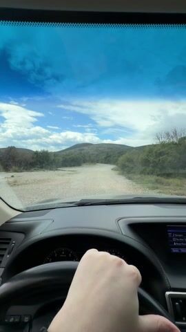 A first-person perspective video shows someone driving on a dirt road leading towards a hill. The sky is blue with scattered clouds. The dashboard of the vehicle is visible, including the steering wheel, speedometer, and infotainment screen. The driver’s left hand, adorned with a black beaded bracelet, is visible on the steering wheel. The road ahead is unpaved and surrounded by sparse greenery.
