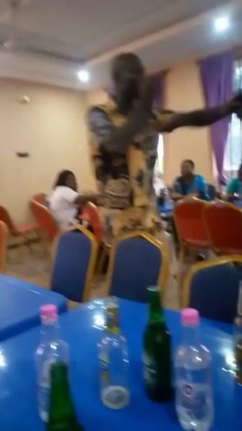 A man in a colorful shirt and khaki pants is dancing at a table filled with various bottles of beverages. He appears to be pouring beer into his mouth directly from a bottle, while moving to the music. A person wearing a pink shirt with the number "22" is also nearby, holding a bottle. The scene takes place in what appears to be a banquet hall or event space, with multiple tables and chairs. Other people are visible in the background, some seated and engaged in conversation.  The lighting is artificial, and the room has purple curtains.
