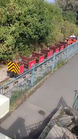 A miniature train is seen from a high angle, moving from right to left on a track. The train is bright orange and black, with a single driver wearing a white shirt. Behind the locomotive are several open carriages, each with wooden benches and red signs. The train is surrounded by lush green trees on the left and a metal fence to the right, separating it from a paved pathway. A concrete structure is visible in the lower left corner.