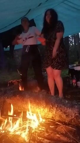 Two individuals are dancing around a campfire under a blue tarp. The person on the left wears a white t-shirt and dark pants, while the person on the right wears a black and white patterned dress. The fire is burning brightly in the foreground. The environment appears to be outdoors, possibly a campsite, with trees visible in the background. The lighting is dim, suggesting it is either dusk or night.