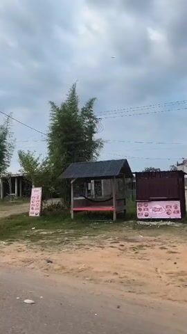 A view of a rural area under an overcast sky, taken from a moving vehicle. The shot shows a roadside scene with sparse vegetation and a mix of buildings, some under construction and others appearing to be abandoned or in disrepair. A weathered sign reading "SHIMIZO UD PUTRA JAYA ABADI" is visible. A small, open-sided structure with a hammock hangs in the background. The road is paved but shows signs of wear and tear. No people are clearly visible in the video.