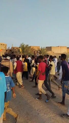Here is a description of the video:

The video shows a large group of people walking and milling around in an open, sandy area. Most of the people are young men, but there are people of different ages present. Some people are wearing traditional white robes, while others are wearing casual clothing like t-shirts and jeans. There is a yellow flag in the crowd. The background features low, mud-brick buildings and palm trees, suggesting a rural or desert environment. The sky is light blue and the light suggests it is either dawn or dusk. The general atmosphere seems lively and festive.
