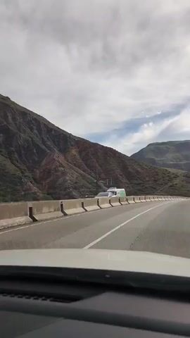 A first-person perspective video shows a car driving on a highway with a mountainous landscape. The highway has concrete barriers separating the lanes. The mountains are covered in sparse vegetation, with some rocky, eroded sections. The sky is cloudy. Several vehicles are visible on the road, including a white van with green markings and a silver SUV. The video captures the winding road and the vastness of the natural scenery.