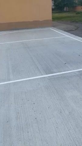 A video shows a tennis ball bouncing on a concrete court, which has white lines. The court is adjacent to a light-brown building. The camera remains static during the video. The video is shot outdoors in daylight.