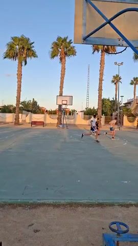 A basketball court with a chain-link fence around it is seen in the video. There are several palm trees on the left side of the court, as well as some buildings in the background. In the foreground, there is a basketball hoop with a blue pole. Three people are visible on the court, one of them is playing basketball and the other two are standing. The video is taken at sunset, and the sky is clear and blue.